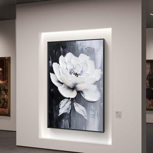 Puede incluir: Pintura floral enmarcada que representa una peonía blanca sobre un fondo texturizado en blanco y negro. La obra de arte se exhibe en una galería, iluminada por iluminación empotrada, agregando un toque de elegancia al espacio.