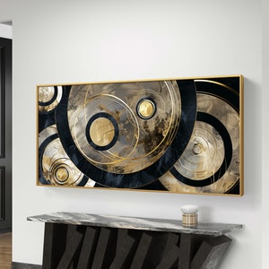 以下が含まれることがあります： ベージュの背景に、金色と黒色の同心円が描かれた抽象的なアートプリント。プリントは薄い金色のフレームで額装されています。