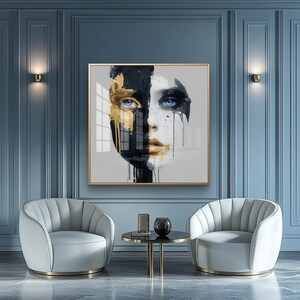 Könnte beinhalten: Ein gerahmter Kunstdruck mit einem geteilten Gesichtsdesign. Eine Seite ist schwarz und gold, die andere weiß mit schwarzen Tropfen. Das Motiv hat blaue Augen und goldene Lippen. Das Kunstwerk hängt über zwei weißen Sesseln und einem kleinen Tisch.