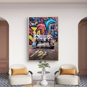 Puede incluir: Una impresión enmarcada con un diseño de estilo grafiti en un mando de videojuegos. El mando está pintado con colores vibrantes y colocado contra un fondo de pared de grafiti colorido. La impresión se exhibe en una habitación con dos sillones y una mesa pequeña.