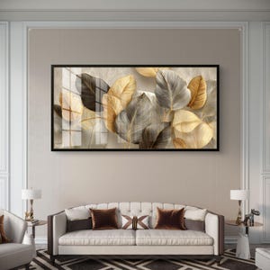 Op de afbeelding: Een ingelijst kunstwerk met gestileerde gouden, grijze en bruine bladeren. Het kunstwerk wordt weergegeven boven een witte bank met bruine kussens. De kamer heeft muren in neutrale tinten en bijzettafels met lampen.