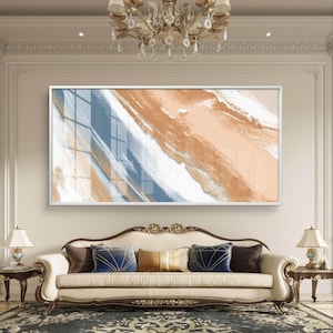 Puede incluir: Gran obra de arte abstracta enmarcada con un marco blanco. La obra presenta una mezcla de pinceladas melocotón, blancas y azules. La pintura cuelga sobre un sofá color crema con cojines decorativos.