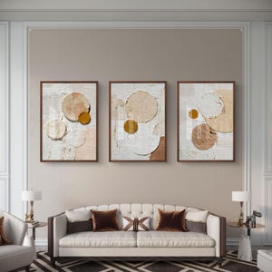 Peut inclure: Trois peintures abstraites encadrées avec un fond blanc texturé et des cercles superposés beiges, bronzés et dorés. Les peintures sont exposées au-dessus d'un canapé blanc avec des coussins bruns.