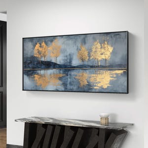 Puede incluir: Pintura abstracta de paisaje con árboles de pan de oro reflejados en el agua. La obra de arte tiene un fondo azul oscuro y gris con una línea horizontal dorada. La pintura está enmarcada en un marco negro y cuelga sobre una consola moderna.