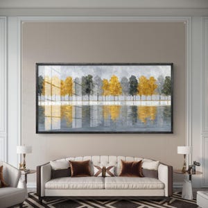Puede incluir: Una pintura de paisaje enmarcada representa una fila de árboles en tonos dorados, grises y verdes reflejados en el agua. La obra de arte se exhibe sobre un sofá blanco con cojines marrones. La habitación tiene paredes de tonos neutros y una alfombra estampada. El cuadro mide unos 150 cm de ancho.