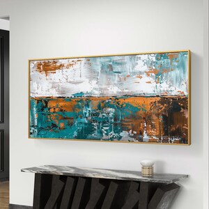 Puede incluir: Pintura abstracta en un marco dorado, con un diseño texturizado en tonos blanco, turquesa y naranja. La obra de arte cuelga sobre una consola moderna con tapa de mármol y base negra geométrica. Una pequeña lámpara está sobre la mesa.