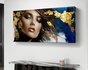 Peut inclure: Une œuvre d'art sur toile rectangulaire représentant un portrait de femme avec des accents de feuilles d'or. La femme a les yeux bleus et du maquillage doré. Le fond présente des coups de pinceau bleus et blancs. L'œuvre est affichée au-dessus d'une console noire et grise.