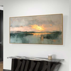 Può includere: Un dipinto astratto orizzontale di paesaggio con una cornice dorata. Il dipinto presenta una tavolozza di colori tenui di blu, verdi e rosa. Il cielo è azzurro chiaro con nuvole vaporose, e l'orizzonte è una linea di colline verdi. L'acqua è blu scuro, e il sole tramonta in lontananza, proiettando un bagliore dorato sulla scena.