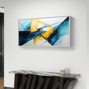 May include: Abstract art piece in a white frame, featuring a dynamic composition of blue, yellow, and black geometric shapes. The artwork hangs above a modern console table with a marble top and a dark, angular base. A small decorative lamp sits on the table.
