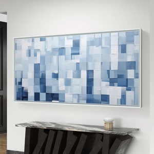 Op de afbeelding: Een groot abstract schilderij in blauw en wit. Het schilderij is ingelijst in een wit frame en hangt aan een witte muur. Een zwart-wit marmeren consoletafel met een geometrisch ontwerp staat op de voorgrond.