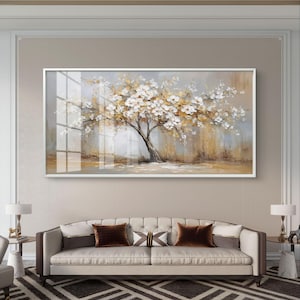 May include: A large framed painting of a tree with white and gold blossoms. The artwork hangs above a beige sofa with brown throw pillows. The room has a neutral color palette with a patterned rug and side tables with lamps.