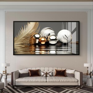 May include: A framed print showing a gold feather and metallic spheres in gold, silver, and white with a marble design. The spheres reflect in a rippled surface. The artwork hangs above a beige sofa with brown cushions, creating a modern interior design.