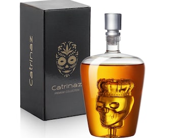 May include: A clear glass decanter with a skull design and a silver stopper, filled with amber liquid. The decanter is next to a black box with the name "Catrinaz" and a skull design. The box reads "Catrinaz Premium Collection."