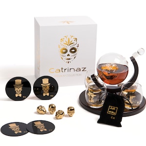 Könnte beinhalten: Ein Dekanter-Set aus schwarzem und goldenem Totenkopf mit vier passenden Gläsern. Der Dekanter hat einen runden Körper und einen gebogenen Ausguss. Die Gläser sind kurz und rund. Das Set wird mit einem schwarzen Samtbeutel geliefert, auf dem in Gold "Just enjoy" steht. Das Set ist in einer weißen Box mit einem goldenen Totenkopfdesign und den Worten "Catrinaz Premium Collection" verpackt.