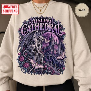 May include: A sand-colored crewneck sweatshirt featuring a graphic print of a gargoyle perched on a stone pedestal in front of a cathedral. The design includes purple roses, moths, and the text "AISLING CATHEDRAL" in a stylized font.