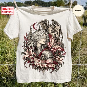 May include: An ivory t-shirt featuring a graphic print of a blindfolded knight in armor, a gargoyle, a large moth, lilies, and a crescent moon. A banner at the bottom displays the text, THE KNIGHT AND THE MOTH, in a dark red font.