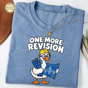 May include: A blue crew neck t-shirt featuring a graphic of a white goose wearing a yellow hard hat and black glasses. The goose holds blue rolled blueprints and a flat blueprint of a bridge. The text ONE MORE REVISION is printed above.