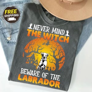 May include: A charcoal gray t-shirt featuring a graphic of a Labrador dog centered in front of a large orange moon. The design includes silhouettes of a haunted house, bare trees, and a flying witch, with the text "NEVER MIND THE WITCH BEWARE OF THE LABRADOR" printed in white and orange.