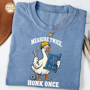 May include: A blue crew neck t-shirt featuring a graphic of a white goose wearing a yellow hard hat and glasses. The goose holds a caliper and a blue blueprint. The text reads MEASURE TWICE. HONK ONCE. The shirt has a faded, vintage-style finish.