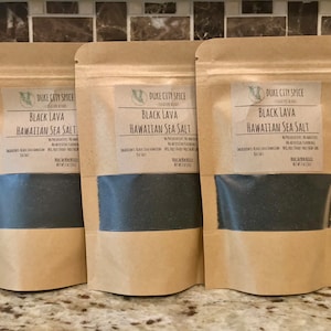 May include: Three brown paper bags filled with black lava Hawaiian sea salt. Each bag has a white label with the text "DUKE CITY SPICE" and "BLACK LAVA HAWAIIAN SEA SALT".