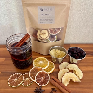 May include: A brown paper bag with a label that reads "Duke City Spice" and "Sangria Wine Kit". The bag contains dried fruit, spices, and citrus slices. There is a glass jar filled with red wine, cinnamon sticks, and a star anise. There are also small bowls filled with dried fruit and citrus slices.