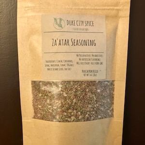 May include: A brown paper pouch with a white label that reads "DUKE CITY SPICE - SIGNATURE BLENDS - ZA'ATAR SEASONING". The label lists the ingredients: cumin, coriander, thyme, marjoram, sumac, organic white sesame seeds, and sea salt. It also states that the seasoning is made in New Mexico and is free of preservatives, additives, artificial flavorings, MSG, dairy, and GMOs. The pouch contains a brown, green, and white spice blend. The pouch is labeled with a net weight of 1 oz (28g).