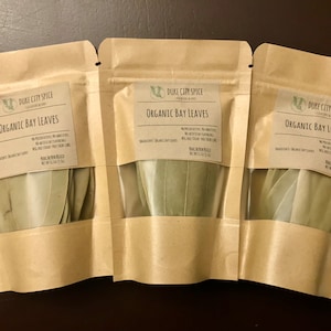May include: Three brown paper bags with clear windows, each containing a bundle of dried organic bay leaves. The bags are labeled "DUKE CITY SPICE" and "ORGANIC BAY LEAVES".