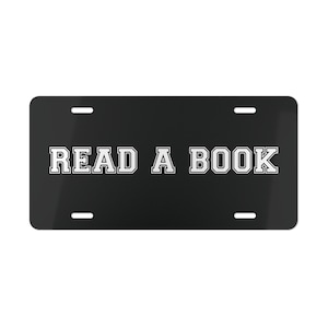 Op de afbeelding: Zwarte nummerplaat met de tekst "READ A BOOK" in het wit, in een lettertype in collegestijl. De plaat heeft vier montagegaten en een glanzende afwerking.