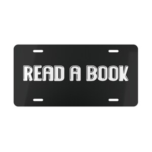 Op de afbeelding: Zwarte nummerplaat met de tekst "READ A BOOK" in vette, witte, omlijnde letters. De plaat heeft vier montagegaten. De achtergrond is effen zwart.