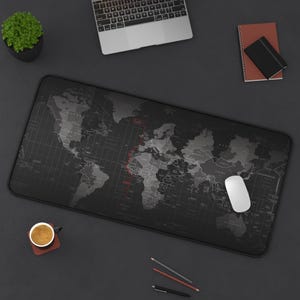 May include: A black desk mat with a world map design, showing gray and white continents and countries. A white computer mouse is on the mat. Other items on the desk include a laptop, a small plant, a notebook, and a cup of coffee.