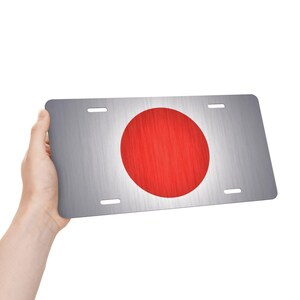 Puede incluir: Placa de matrícula rectangular con acabado de metal cepillado. La placa presenta un gran círculo rojo en el centro, que representa la bandera japonesa. La placa tiene cuatro orificios de montaje y está sujeta por una mano.