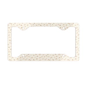 May include: A rectangular license plate frame featuring a cream-colored background with a repeating pattern of small, light brown floral blossoms and tiny dots. The frame has four circular mounting holes, one in each corner, for secure attachment.