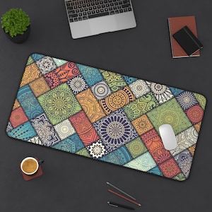 May include: A rectangular desk mat with a colorful patchwork design. The mat features a variety of geometric patterns in shades of blue, green, orange, and yellow. A white computer mouse rests on the mat, alongside a laptop and a cup of coffee.