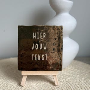 Op de afbeelding: Een klein, vierkant, goudkleurig bord met de woorden "HIER JOUW TEKST" in witte letters, weergegeven op een miniatuur houten ezel. Een witte, abstracte vaas staat op de achtergrond. Het bord heeft een textuur.