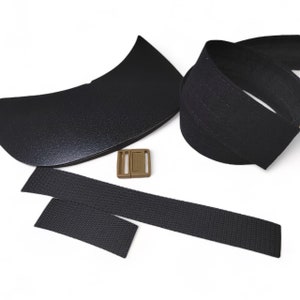 Puede incluir: Un conjunto de correas de tela negra y una hebilla de plástico marrón. Las correas son tejidas y tienen una superficie texturizada. La hebilla es rectangular con dos ranuras.
