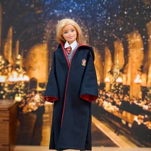 May include: A blonde doll wearing a black robe with red trim and a Hogwarts house crest. The doll is standing on a wooden surface with a blurred background of a room with Christmas decorations.