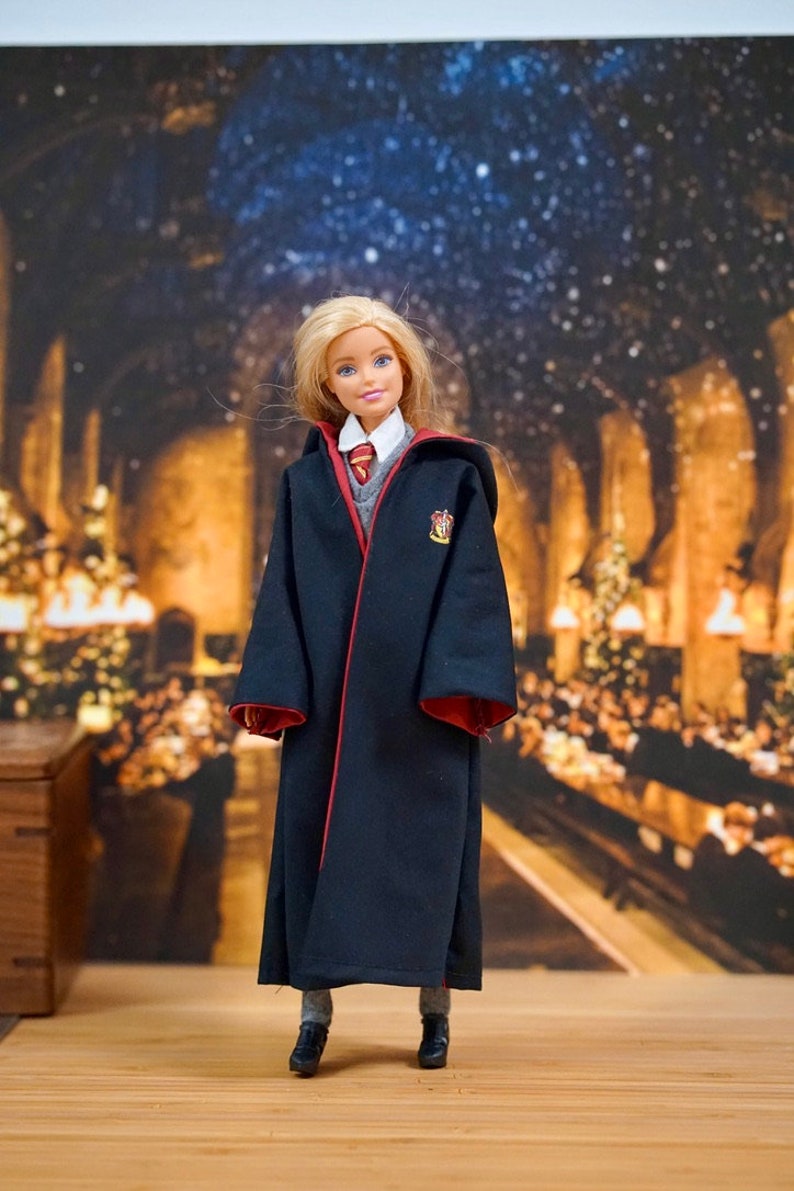 May include: A blonde doll wearing a black robe with red trim and a Hogwarts house crest. The doll is standing on a wooden surface with a blurred background of a room with Christmas decorations.