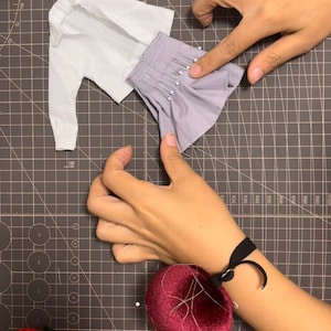 May include: A white shirt and a purple skirt are pinned together on a gray cutting mat. A red pincushion is on the right side of the image.