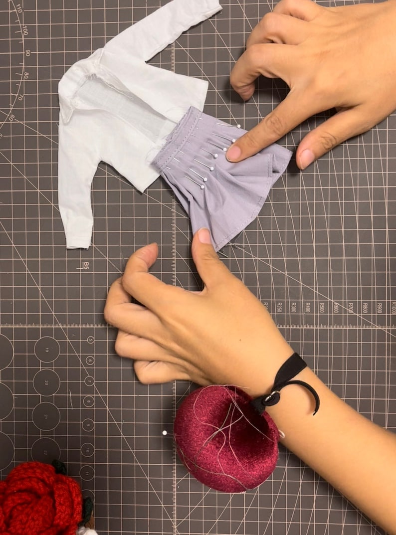 May include: A white shirt and a purple skirt are pinned together on a gray cutting mat. A red pincushion is on the right side of the image.