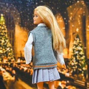 May include: A blonde doll wearing a grey vest with a red and yellow stripe, a white shirt, a light blue pleated skirt, and grey socks. The doll is standing in front of a blurry background of a Christmas scene with lights and trees.