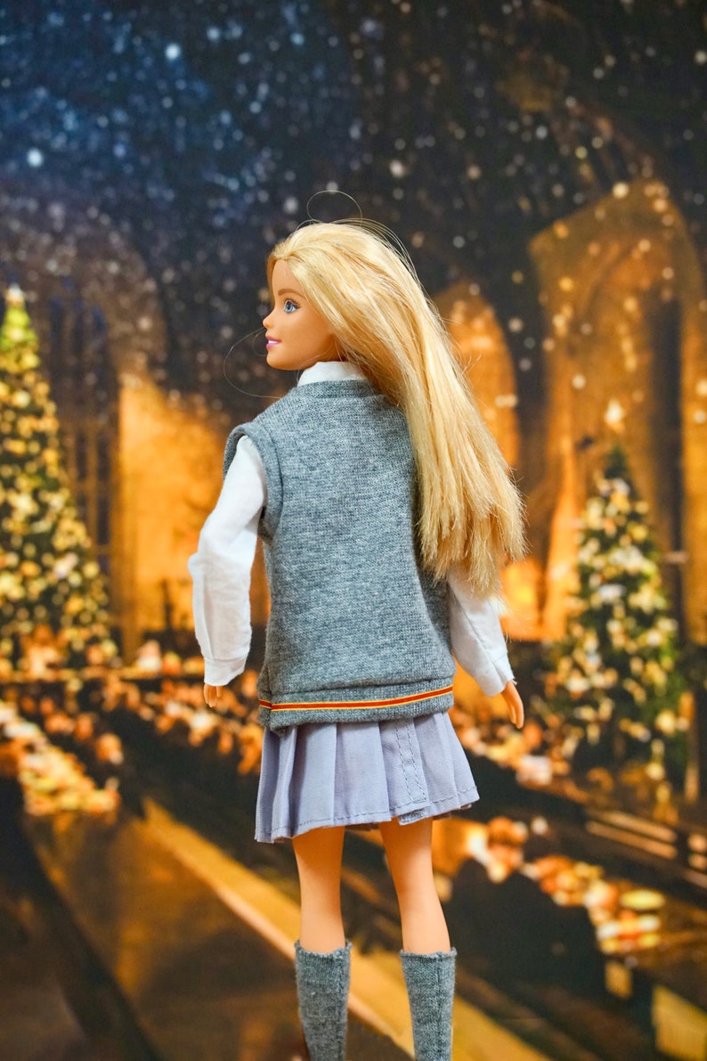 May include: A blonde doll wearing a grey vest with a red and yellow stripe, a white shirt, a light blue pleated skirt, and grey socks. The doll is standing in front of a blurry background of a Christmas scene with lights and trees.