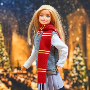 May include: A blonde doll wearing a grey vest, a red and gold scarf, and a pleated skirt. The doll is standing in front of a blurry background of a room with Christmas decorations.
