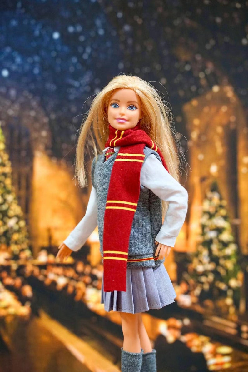 May include: A blonde doll wearing a grey vest, a red and gold scarf, and a pleated skirt. The doll is standing in front of a blurry background of a room with Christmas decorations.