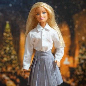 May include: A blonde doll wearing a white button-down shirt and a gray pleated skirt. The doll is standing in front of a blurred background of Christmas lights.
