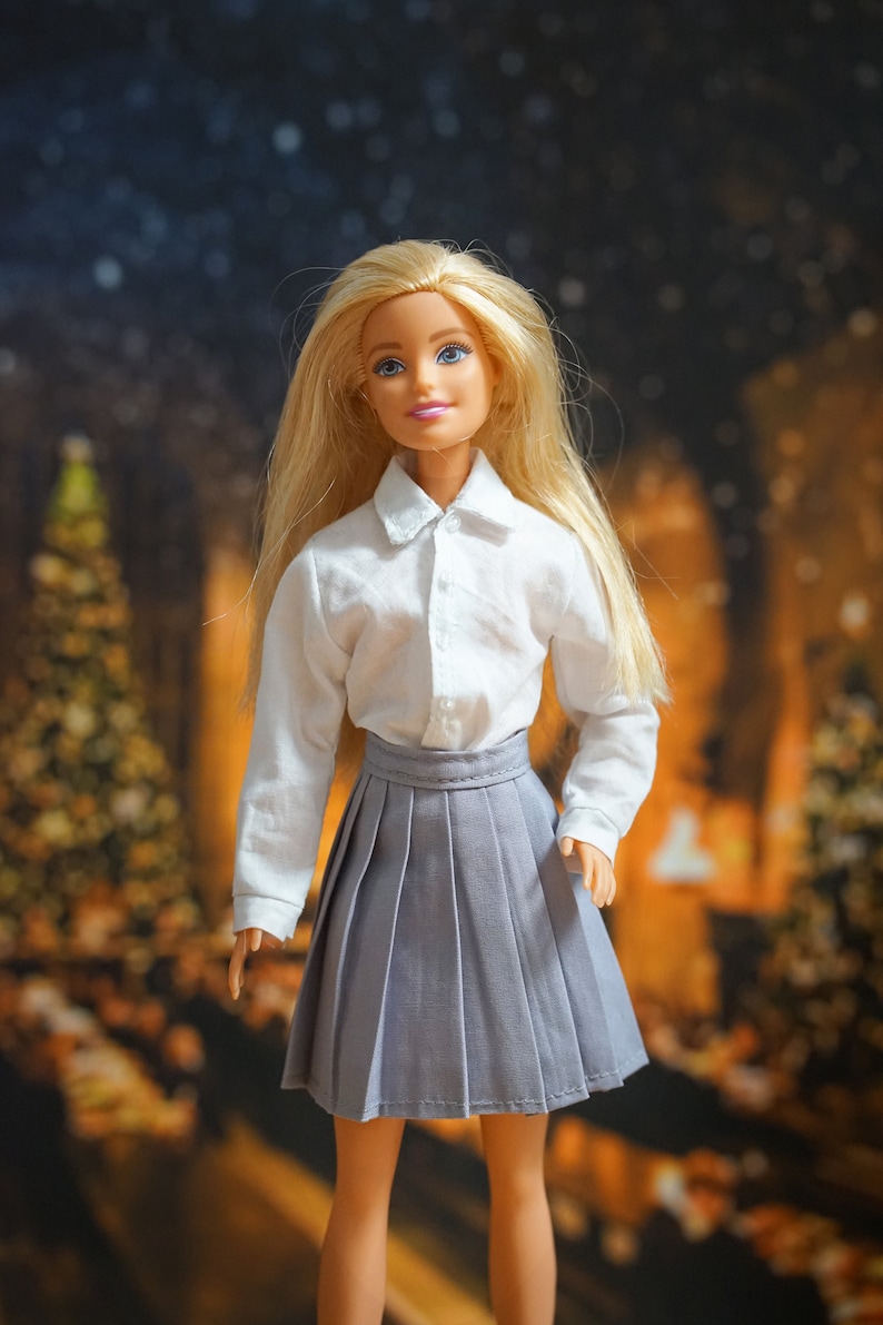 May include: A blonde doll wearing a white button-down shirt and a gray pleated skirt. The doll is standing in front of a blurred background of Christmas lights.