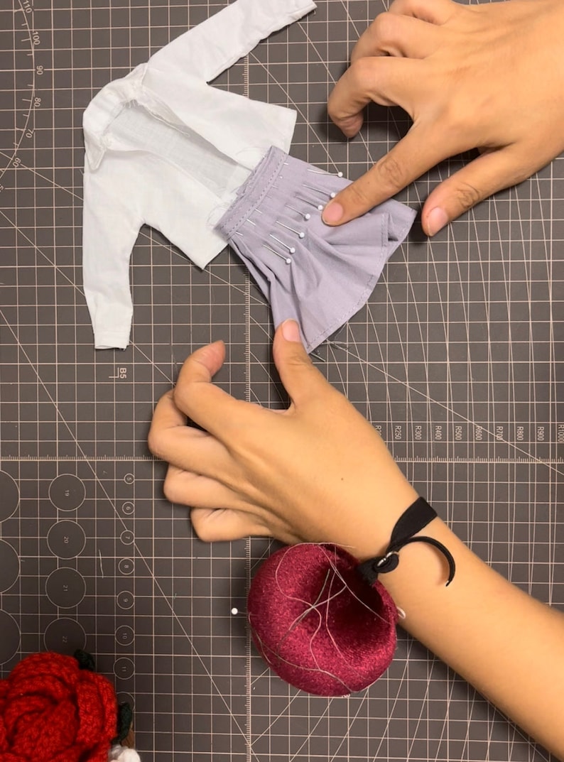 May include: A close-up of a person pinning a light purple skirt to a white shirt on a gray cutting mat. The person is wearing a black wristband with a red pincushion attached.