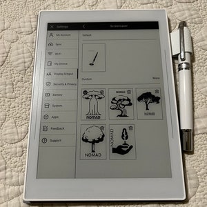 Può includere: Un tablet digitale bianco che mostra il menu delle impostazioni, con opzioni per account, Wi-Fi e display. Lo schermo mostra opzioni di screensaver personalizzate con icone di alberi e la parola "NOMAD". Una penna stilo bianca è a destra.