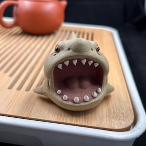 May include: A ceramic shark figurine with an open mouth, displaying white teeth and a pink interior. The shark is light brown with small, dark eyes. It sits on a wooden tray, with a terracotta teapot in the background.