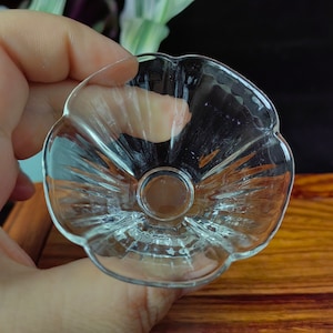 May include: A clear, small glass bowl with a flower-like shape. The bowl has a circular base and a scalloped rim. The glass is transparent, allowing light to pass through. The bowl is held by a hand, with a wooden surface in the background.