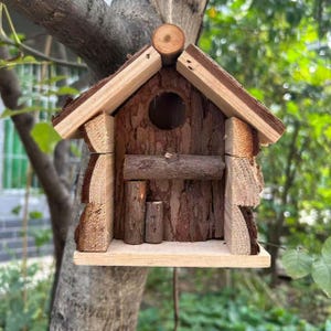 Puede incluir: Una pajarera rústica de madera con exterior de corteza natural y un orificio de entrada redondo. La pajarera cuenta con una pequeña percha y un tejado inclinado, diseñado para dar cobijo a los pájaros pequeños. Cuelga de una rama.